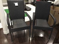 Two charcoal grey poly-rattan patio chairs side by side on dark wood floor, one chair missing two metal decorative feet but still functioning