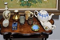 Overall lot showing assortment of vases, teapots, cups, decorative dish, and inkwell arranged on wooden table with framed painting backdrop.