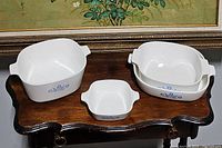All four Corning Ware dishes displayed together on wooden table showing variety of sizes and blue cornflower decals.