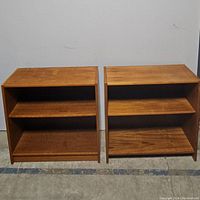 Front view of two wooden bookshelves side by side, showing two shelves each and wood grain finish. Both show some surface wear.