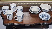 Five white mugs with blue floral pattern arranged on wooden table along with small bowls and plates