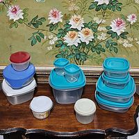 All plastic containers arranged in three stacks on wooden surface with floral painting background. Containers are clear plastic with blue, white, and red lids.