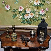 Wide view showing seven decorative items on wooden table including candlesticks, jar, elephant figurine, lantern, and silver bowls with floral painting backdrop.