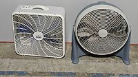 Two floor fans on the ground against a wall: one square white box fan and one round gray and white floor fan.