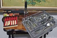 Complete view of table with flatware assortment including boxed steak knives, metal silverware tray with forks, spoons, knives, and serving utensils.