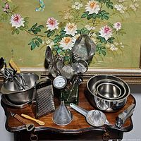 Photo showing various metal kitchen utensils clustered together including graters, ladles, potato masher, meat tenderizer, citrus juicer, funnel, and bottle opener on a wooden surface with floral wallpaper background.