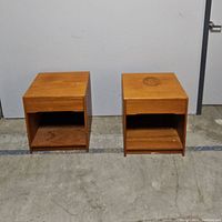 Front view of two wooden side tables showing drawers and open shelves beneath, sitting on concrete floor against a white wall.