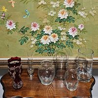 Seven glass vases placed on wooden table with floral picture background: five clear glass vases of varying shapes and sizes, one ruby red glass vase with flared neck, one ruby red glass vase with cut decorative designs and scalloped rim.