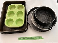 Photo showing nine pieces of bakeware including muffin pan, baking sheet, springform pan, tart pan, and round cake pans on white surface