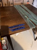 Photo showing the top of a dark brown wooden bar height table with two separate leaves strapped on top, along with blue tape labeling the table and its measurements (36.5" x 36.5" x 35.5").