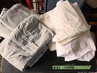 Photo of assorted bed linen pieces in white and gray colors folded and stacked on a black surface with a green label reading '#11 LINEN'.