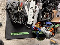 Top-down view of a collection of cables, power bars, tie-down straps, and battery clamps placed on a black table surface with a green tape label reading '#21 Cables & Cords'.