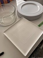 Photo showing one square white ceramic plate with subtle geometric pattern, stacked white round charger plates, and a clear glass bowl.