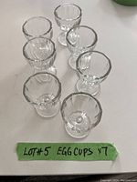 Photo of seven clear glass egg cups arranged on a white surface, showing their fluted design and round bases.