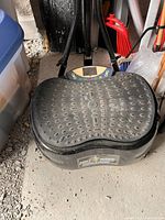 Full view of the black full body vibration machine platform showing the textured rubber surface and the control panel; machine surrounded by household items indicating storage conditions.