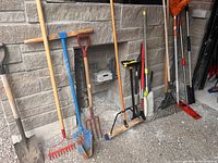 Photo shows multiple garden tools including shovels, rakes, pitchforks, broom, and brushes against a stone wall.