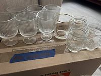 Photo of nine assorted glassware pieces on a cardboard box, six ribbed stemmed glasses, one tumbler with gold rim and three smaller clear tumblers.