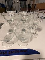 Photo showing assortment of 8 clear glassware items on a white surface including two large wine glasses, two cocktail glasses, two brandy snifters, and two smaller patterned wine glasses.