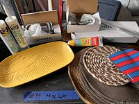 Photo of a yellow corn tray, three woven placemats including one with stripes, adhesive tubes, and boxed Moen bathroom accessories.