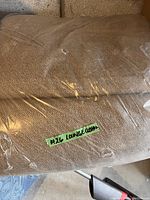 Photo of beige outdoor lounge chair cushion wrapped in plastic, showing fabric texture and a small green label stating '#26 LOUNGE CUSHION'.