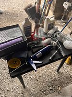 Assortment of paint accessories laid on a black folding table including rollers, trays, brushes, brushes in jar, caulking gun, and liquids