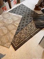 Three area rugs on floor showing one small dark grey/black patterned accent rug, a medium black and white geometric patterned rug, and a larger beige and brown medallion design rug.