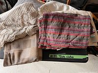 Four folded textile pieces laid on a black surface including a beige and brown checkered tablecloth, a beige lace crocheted cloth, a plain beige fabric cloth, and a rectangular striped table runner in shades of red, purple, and brown.