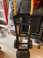 Photo showing a black metal hand truck trolley with yellow handles, large black wheels, and a toe plate, positioned upright on a concrete floor near workshop items.