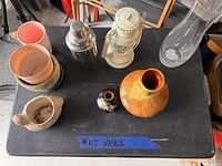 Eight assorted items displayed on a black table including plant pots, a cocktail shaker, an oil lamp, vases, and a mortar with pestle.