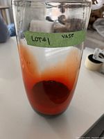 Photo of a tall cylindrical glass vase with a gradient color from red at the base fading to clear at the top. A green tape label is wrapped around the upper part of the vase that reads 'LOT #1 VASE'.