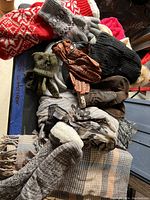 Top-down photo showing assorted winter wear items piled on a flat surface. Includes gloves, knit hats, and scarves in multiple styles and colors.