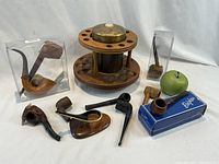 Photo showing twelve vintage pipes including boxed pipe, Calabash pipe with white Meerschaum bowl, bull head pipe, corncob pipe, and various handcrafted wooden pipes. Circular wooden pipe rack with humidor and brass lid, plus single pipe stand with brass accents visible.