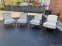 Four mahogany armchairs with ivory cushions arranged outdoors on pavement, wooden fence and brick wall in background