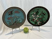 Two cloisonne plates on stands with a green apple for scale.