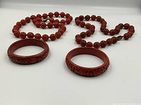 Two sets of round carved cinnabar lacquer bead necklaces with matching carved bangle bracelets arranged on white background.