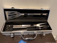 Open aluminum carrying case showing three BBQ tools: slotted spatula, fork, and tongs, each held with black straps on the foam insert.