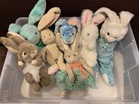 Seven assorted large stuffed bunnies in a plastic bin with various colors and styles, including plush, fabric, and knit textures.