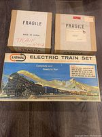 Photo of the original Lionel Electric Train Set box with artwork showing steam locomotive, train cars, and mountainous background, along with two fragile labeled boxes.