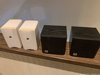 Two pairs of small speakers: two white rectangular JBL TLX Movies Surround speakers and two black cube-shaped Mirage AVS-200 speakers.