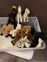 Eight assorted stuffed horse plush toys arranged in a plastic bin, colors include black with yellow, white with pink mane, white with brown spots, and others.