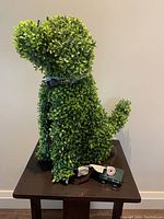 Side view of the topiary dog showing dense artificial green leaves, checkered collar, and light string battery pack below.