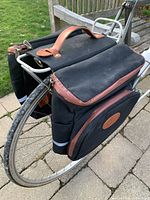 Rear bicycle pannier bags installed on a bike rear rack showing black canvas material, brown leather accents, and side hook attachments.