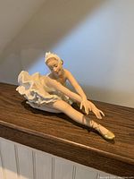 Front and side views of White porcelain bisque ballerina figurine sitting and stretching her leg forward wearing gold pointed ballet shoes.