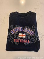 Folded view of the black vintage England Football T-shirt showing raised 'ENGLAND' lettering, England flag, and 'FOOTBALL' text with decorative elements.