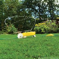 Yellow vintage traveling sprinkler operating on a green lawn, showing the adjustable spray arm spraying water and the spiked rear wheels designed to move along the hose path.