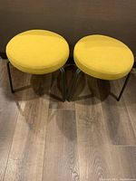 Pair of two round ottoman stools upholstered in yellow fabric with metal legs on wooden floor.