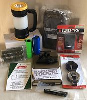 Photo of lantern, flashlights, multi-tool, knives, belts, and survival gear in original packaging and loose, showing variety of camping and outdoor gear.