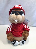 Front view of plush beaver wearing sunglasses, red jacket with white stripes, red and white hat, black and white shoes, sitting upright on white surface.