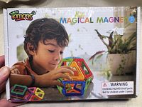 Front view of sealed Tomons Magical Magnet building blocks set showing child playing with colorful magnetic pieces in various geometric shapes.