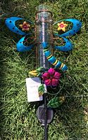 Image shows a colorful butterfly-shaped garden stake with attached rain gauge in a garden setting on grass.